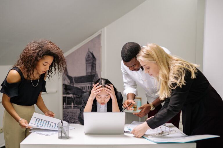 Frau am Laptop schlägt die Hände über dem Kopf zusammen, weil sich die Kollegen um sie herum versammeln. Titelbild zu 'Brauchst du wirklich ein Meeting'
